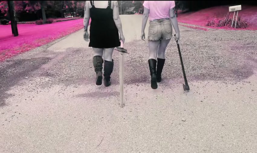 Two women walk down a street, one is holding a sword and the other an axe. The grass is pink.