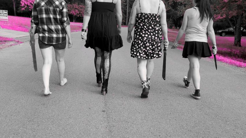 A still from the "Walking With Weapons" music video. Four women walk down a street, each holding a weapon. From left to right: a plank of wood with nails in it; a set of keys between fingers; a machete; a stun gun and a bowie knife. The image is mostly black and white, but the grass framing the women is bright pink where it should be green.